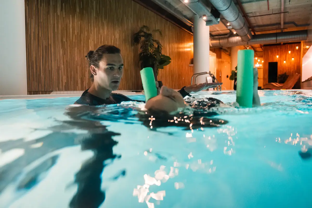 A coach stands close beside a swimmer practising a supported float with two foam noodles